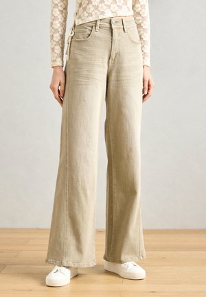 Bežové nohavice s širokými nohavicami a vysokým pásom, s prešitím a zapínaním na gombík. Nosené s bielymi teniskami pre kontrast.