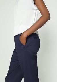 Personne portant un haut blanc sans manches et un pantalon bleu marine avec une main dans la poche avant sur un fond uni.