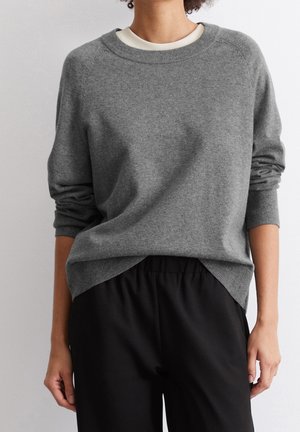 Osoba ubrana w szary sweter z długim rękawem na białą koszulę, w czarnych luźnych spodniach, stojąca na jednolitym jasnym tle.