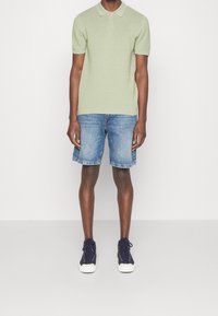 Jasnozielona koszulka polo z krótkim rękawem, niebieskie szorty dżinsowe z podwiniętymi mankietami oraz ciemnogranatowe trampki z białymi podeszwami. Prosty wzór, swobodny styl.