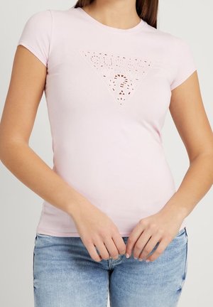 Kobieta w jasnoróżowej dopasowanej koszulce z wyciętym trójkątnym logo Guess i jasnoniebieskich dżinsach, ręce luźno złączone na wysokości talii.