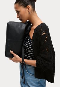Svart läderclutch hålls nära; strukturerad stickad svart cardigan med öppna detaljer; randig svart och vit topp; lösa blå denimjeans.