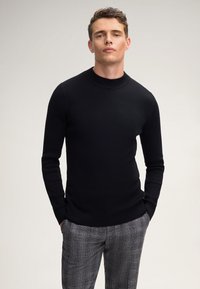 Uomo che indossa un maglione nero a coste aderente e pantaloni grigi a quadri, in piedi con le mani in tasca su uno sfondo chiaro e uniforme.