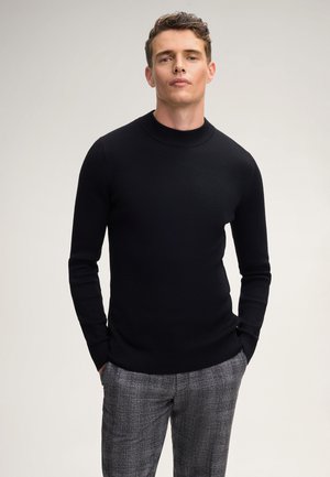 Mann in einem enganliegenden schwarzen Rippenpullover und grauen Karohosen, der mit den Händen in den Taschen vor einem schlichten hellen Hintergrund steht.