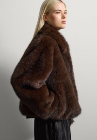 Chaqueta de piel de imitación marrón con un cuello ancho, corte holgado y bolsillos laterales. La textura es densa y suave, proporcionando calidez y comodidad.