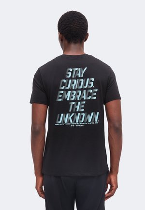 Uomo con capelli corti e ricci che indossa una maglietta nera con testo blu "STAY CURDUS. EMBRACE THE UNKNDWN." sulla schiena, in piedi di spalle.