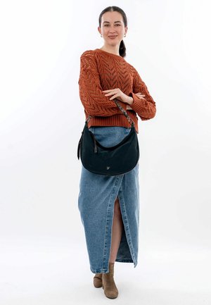 Borsa hobo nera con forma arrotondata e tracolla marrone a contrasto. Presenta una superficie liscia e un dettaglio con logo dorato; indossata con un maglione di lana marrone e una gonna di jeans.