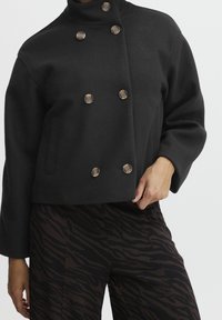 Černá krátká bunda s vysokým límcem, dvouřadým designem a velkými hnědými knoflíky. Spojena s hnědočernými kalhotami se vzorem.