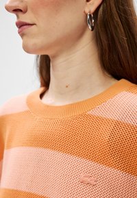 Gestreepte trui in oranje en roze tinten, met een gestructureerde breiwijze, een wijde hals en een klein geborduurd logo op de borst. Zilveren ringoorbellen.