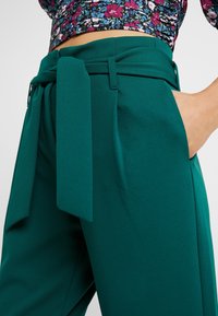 Turkusowe spodnie z wysokim stanem, z materiałowym paskiem w talii, posiadające boczne kieszenie i gładką teksturę. Zbliżenie na detal w okolicy talii.