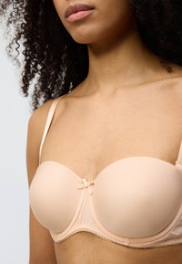 Lys beige balconette-BH med glatt stoff, formpressede kopper, dekorativ sløyfedetalj i midten og justerbare satengstropper.