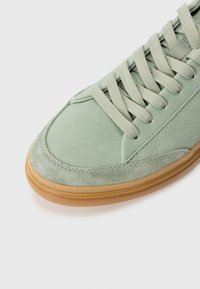 Zapatilla de ante verde con parte superior texturizada, lado perforado y cordones planos. Cuenta con una suela de goma marrón claro.