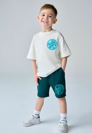 Smilende ung dreng stående med hænderne i lommerne, iført hvid t-shirt og mørkegrønne shorts med matchende cirkulære blå logoer, hvide sneakers.