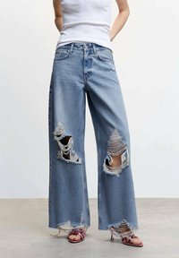 Jean en denim bleu décontractés à jambe large, avec de grandes déchirures et des ourlets effilochés. Associé à un débardeur blanc et des sandales à lanières roses.