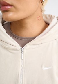 Krémová mikina na zip z měkké tkaniny, s malým bílým logem Nike a tetováním červeného srdce na krku.