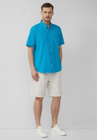 Kurzärmliges türkisfarbenes Hemd mit Knopfleiste und Brusttasche, kombiniert mit hellbeigen, gemusterten Shorts und weißen Turnschuhen. Schlichte und legere Optik.