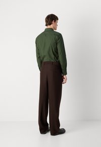 Chemise verte à manches longues avec un col, associée à un pantalon large marron, vue de dos. Tissu lisse, brillance subtile et coupe classique.
