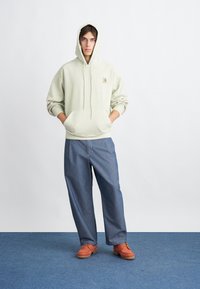 Sudadera verde claro con un bolsillo frontal y un logo, combinada con pantalones anchos de color azul y zapatos naranjas. Textura suave y corte casual.