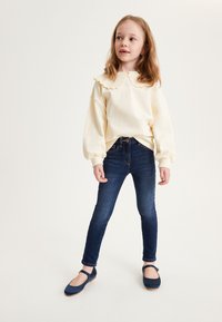 Felpa color crema con colletto a scallop, jeans skinny blu scuro e scarpe Mary Jane navy, con cinturino rotondo e suola piatta.