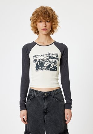 Ribbed crop top with cream body and navy long sleeves. Features a graphic print reading "MUSIC ON WORLD OFF" in black. Paired with dark denim pants.