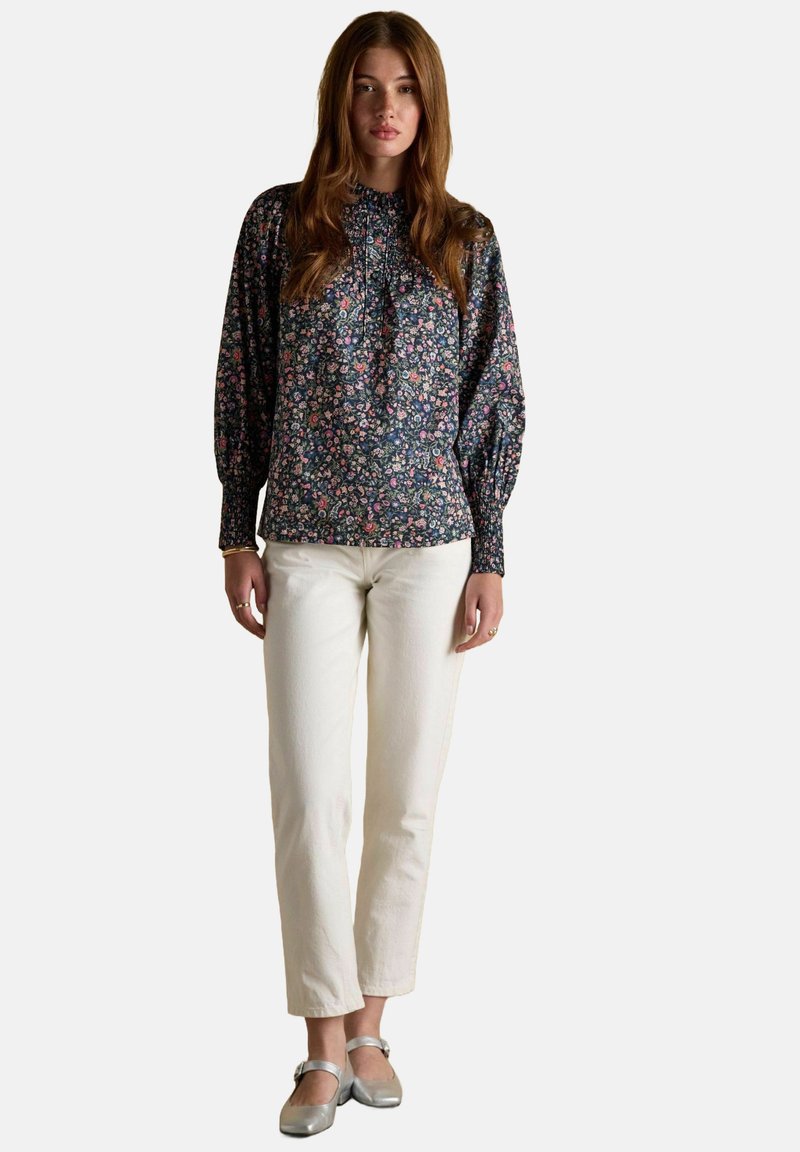 Sieviete, tērpusies ziedu raksta garām piedurknēm blūzē, baltās taisnās biksēs un sudraba Mary Jane kurpēs, stāv pret vienkāršu fona.