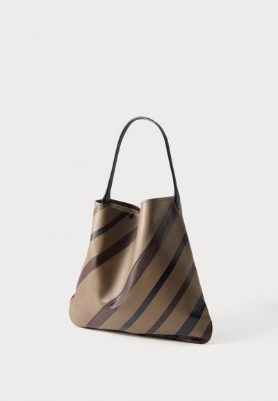 Grand cabas en cuir beige, orné de rayures diagonales foncées et muni d’une longue bandoulière noire, présenté sur un fond clair uni.