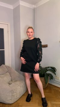 Czarna sukienka z długimi, przezroczystymi, marszczonymi rękawami; prosty, okrągły dekolt. Stylizowana z masywnymi czarnymi butami. W tle naturalna roślina i teksturowane krzesło.