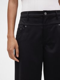 Pantalon noir en tissu lisse, avec une taille ajustée, des poches avant et des accents de boutons métalliques. Ceinture texturée et détails de couture.