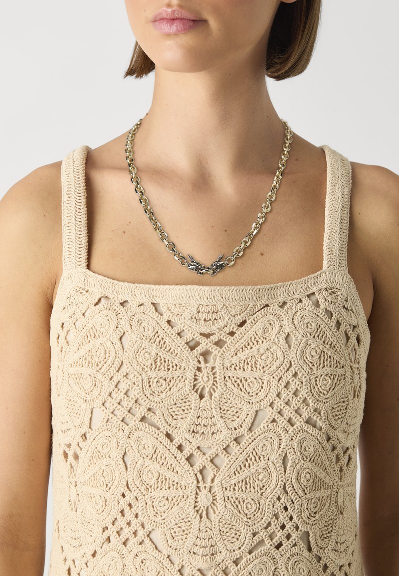 Beige gehäkelte Tanktop mit einem Schmetterlingsmuster, breiten Trägern und einer strukturierten Oberfläche. Eine silberne Kettenhalskette ist ebenfalls sichtbar.