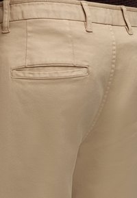 Gros plan d'un pantalon beige montrant une poche arrière avec ouverture passepoilée et des passants de ceinture à la taille.