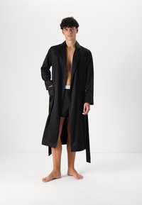 Svart satinlång kåpa med krage, öppen framsida och sidofickor, bärs över svarta shorts, placerad mot en ljus bakgrund.