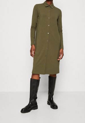 Vrouw die een olijfgroene blousejurk met knopen draagt, gecombineerd met zwarte leren kniehoge laarzen, staand tegen een effen achtergrond.