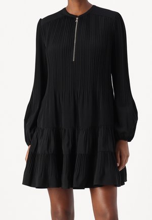 Sort langærmet plisseret minikjole med lynlås foran, lagdelt nederdel og let puffede ærmer, båret af en stående person.