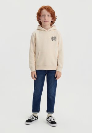 Copil cu păr roșcat stă cu fața spre înainte, purtând un hanorac crem Santa Cruz, blugi albaștri suflecați, șosete crem și adidași alb-negru.