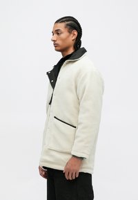 Giacca in pile color crema con colletto nero e bottoni a pressione. Presenta una tasca frontale con rifiniture nere e una vestibilità rilassata. Tessuto testurizzato.