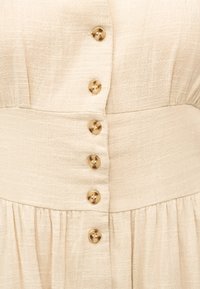 Robe en tissu beige texturé avec six boutons ronds beiges alignés verticalement et un large détail de ceinture.
