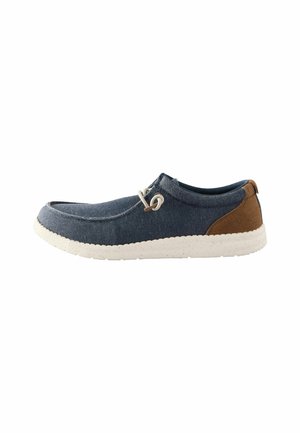 Blue canvas slip-on shoe with a light textured rubber sole. Brown suede accent near the heel. Features a decorative lacing mechanism.