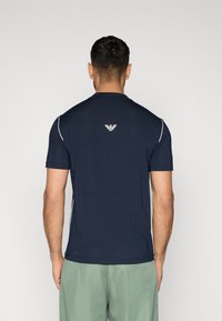 Αθλητική μπλούζα σε ναυτικό μπλε με κοντά μανίκια, λευκές λεπτομέρειες και λογότυπο στην πλάτη. Κατασκευασμένη από ελαφρύ, υφασμάτινο υλικό με υφή.