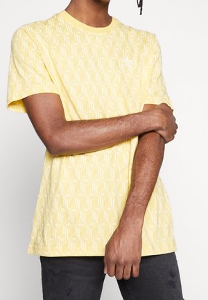 Gelbes T-Shirt mit kurzen Ärmeln, das ein geometrisches Muster in Weiß aufweist, runder Ausschnitt, getragen von einer Person mit verschränkten Armen.