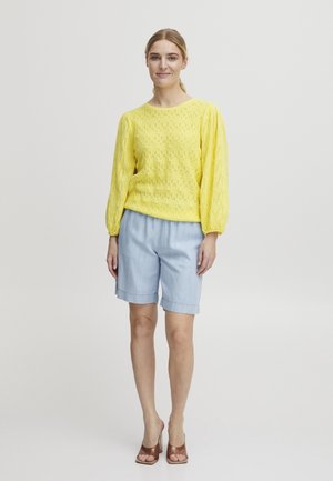 Donna in piedi, indossa una maglia gialla a maniche lunghe testurizzata, pantaloncini celesti fino al ginocchio e tacchi aperti marroni, su sfondo neutro.
