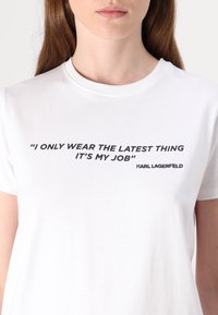 White cotton t-shirt with short sleeves. Front features black text: “I ONLY WEAR THE LATEST THING IT’S MY JOB” and “KARL LAGERFELD.”