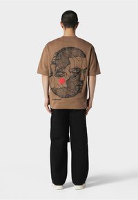 Brun kortærmet t-shirt med et abstrakt sort ansigt designet på ryggen, komplet med en rød detalje i form af en cirkel. Bæres med sorte bukser og hvide sko.
