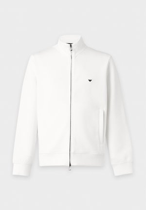 Homme portant une veste zippée blanche, un pantalon noir large et des baskets blanches, debout devant un fond blanc uni.