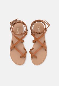 Sandalias de cuero marrón con tiras cruzadas y lazos en el tobillo. Suela plana con bordes cosidos y una textura interior suave. Logo de la marca visible.
