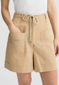 Pantaloni corti beige con vestibilità rilassata, realizzati in tessuto leggero. Presentano tasche frontali, una vita con coulisse e un orlo risvoltato.