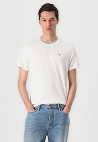 Tricou alb cu mânecă scurtă și guler cu dungi verzi, având un mic logo brodat pe partea stângă a pieptului. Purtați-l cu blugi albaștri deschiși.