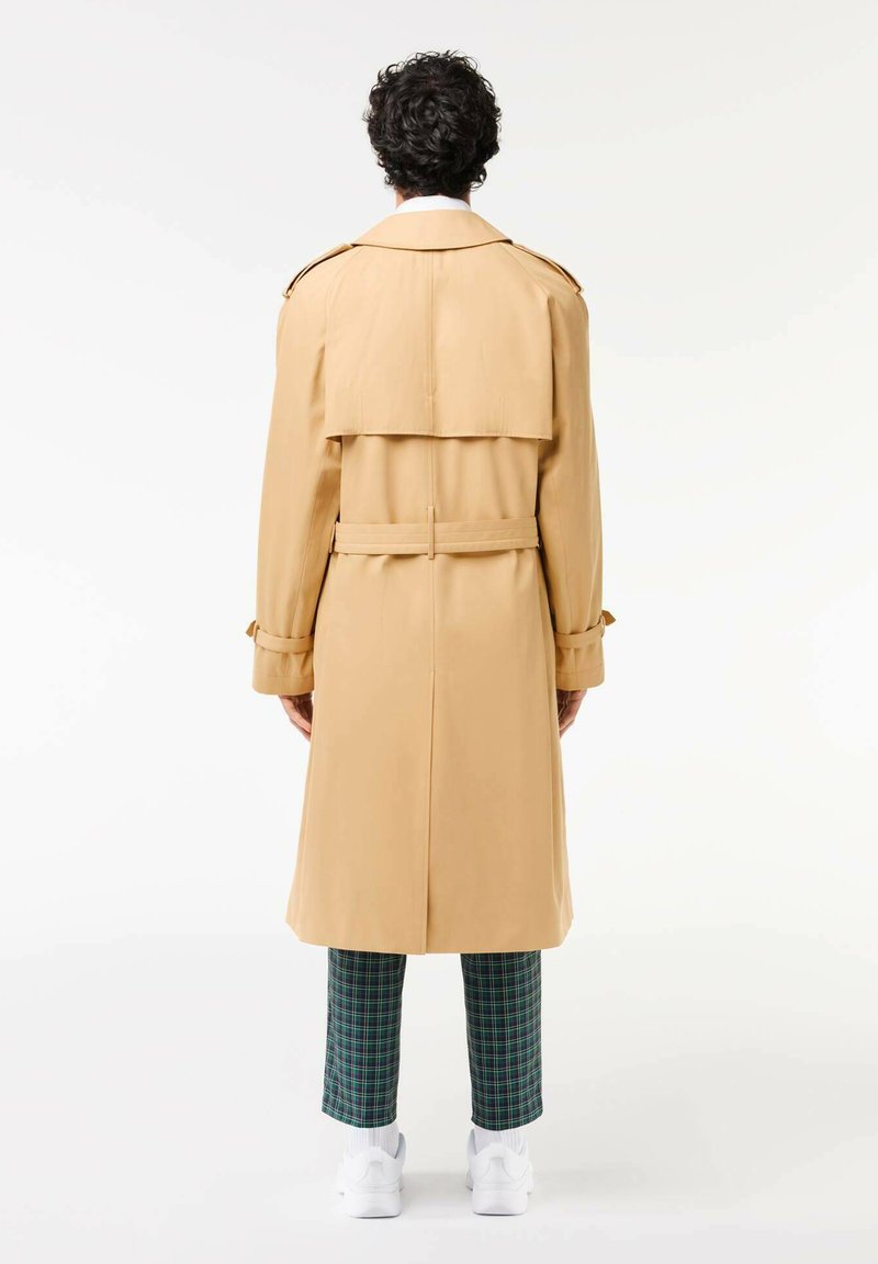 Personne avec des cheveux bouclés portant un trench beige, un pantalon à carreaux vert et bleu, des chaussettes blanches et des baskets blanches, se tenant de dos.