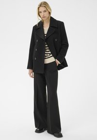 Abrigo negro de doble botonadura con grandes solapas, tela texturizada y detalles de botones, combinado con un suéter de punto a rayas y pantalones de pierna ancha.
