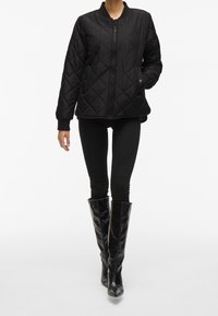 Černá prošívaná bomber bunda se zipem vpředu, žebrovanými manžetami a bočními kapsami, nošená přes černé legíny a černé kozačky sahající ke kolenům.