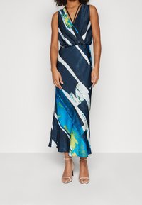 Dlouhá, bezramenná šaty v tmavě modré s abstraktními bílými a akvamarínovými vzory, s překládaným designem a jemnou texturou. Průhledné, páskové podpatky.